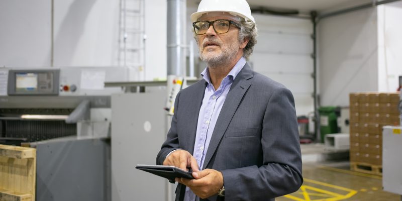 Pensive man in protective helmet holding tablet and looking away. Portrait of Caucasian grey-haired factory CEO in glasses and suit checking project plan. Manufacture and digital technology concept